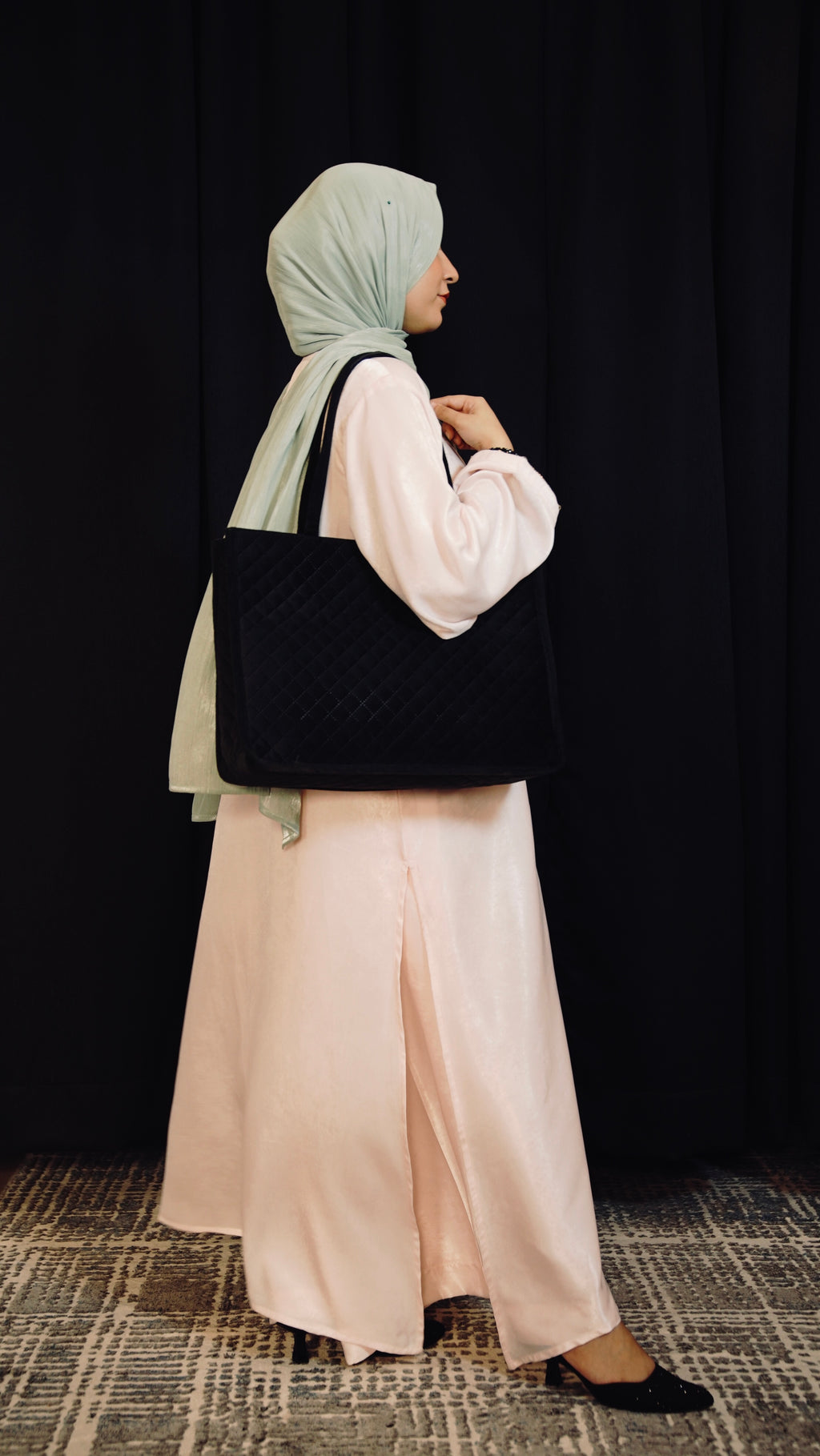 Person wearing a long light pink dress holding a black handbag against a dark background