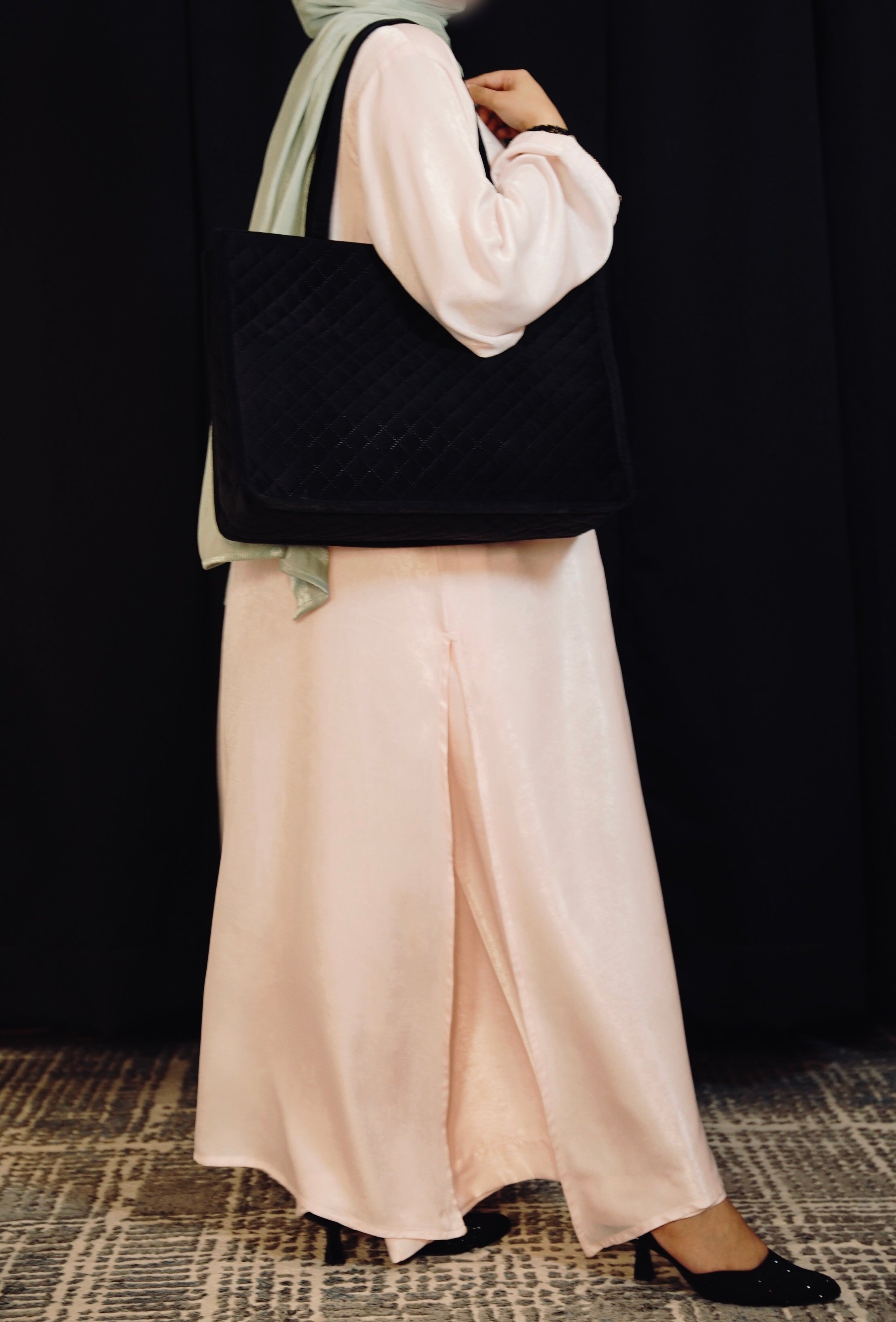 Person wearing a light pink outfit with a black quilted handbag against a dark background